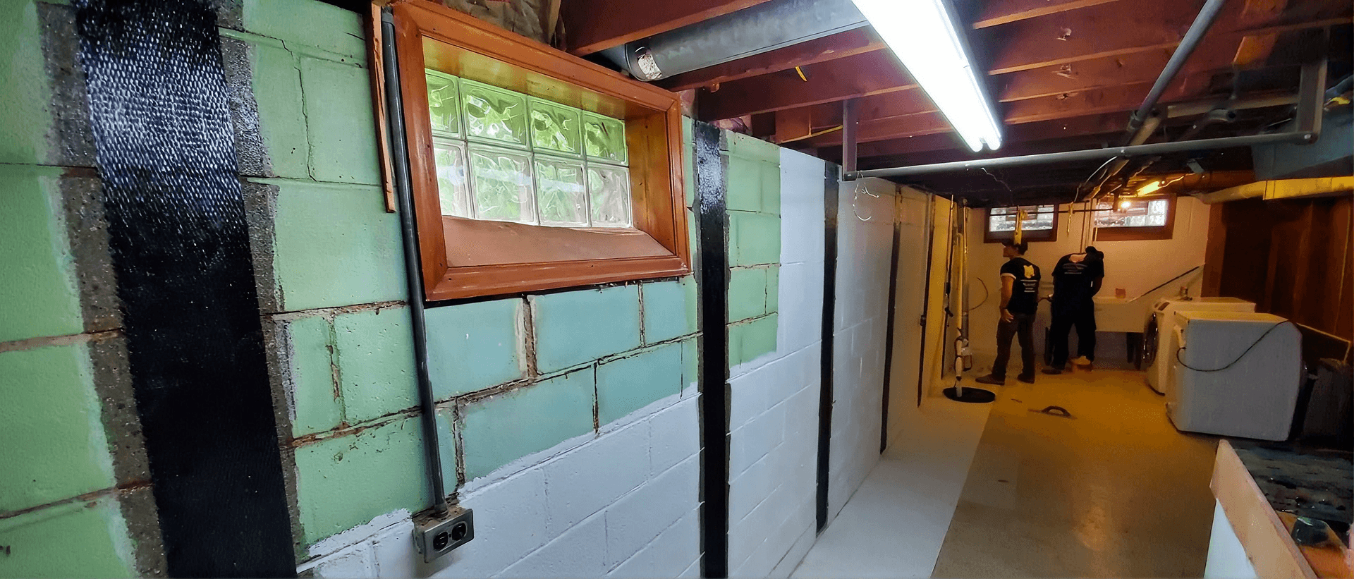 Basement renovation in progress with newly-installed carbon fiber strapping, wooden window frame, fluorescent lighting, and two workers in the background near construction materials.