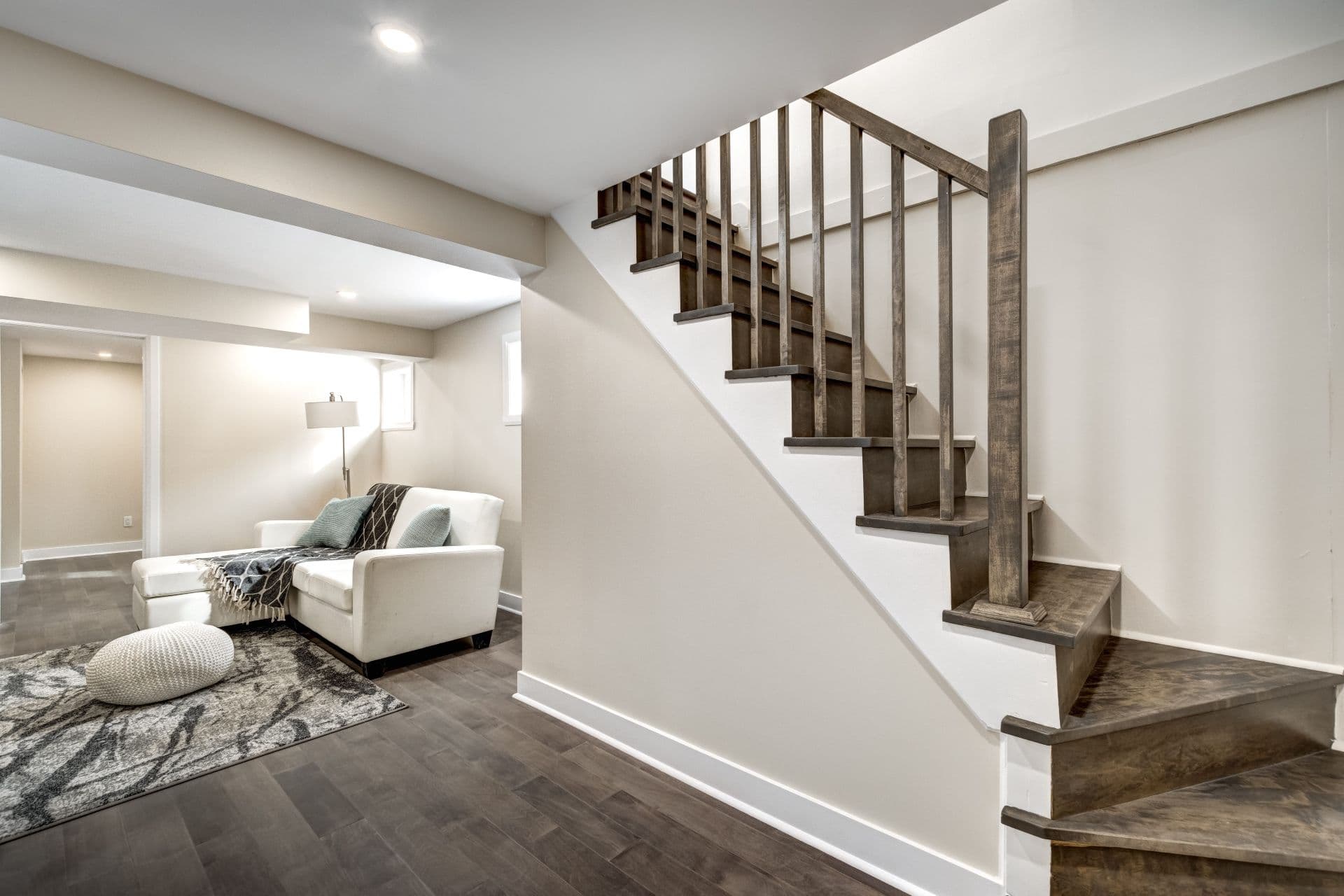 Modern basement living room with cream armchair and wooden staircase with dark railings leading upward.